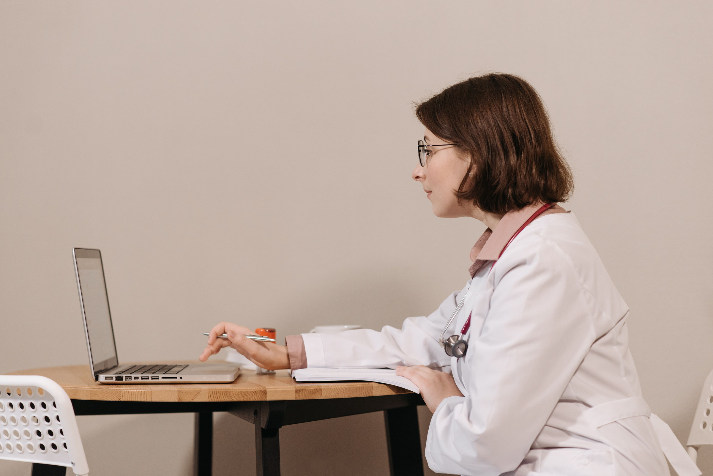 A clinician in a white coat sits in profile at a small wooden table, looking at an open laptop.