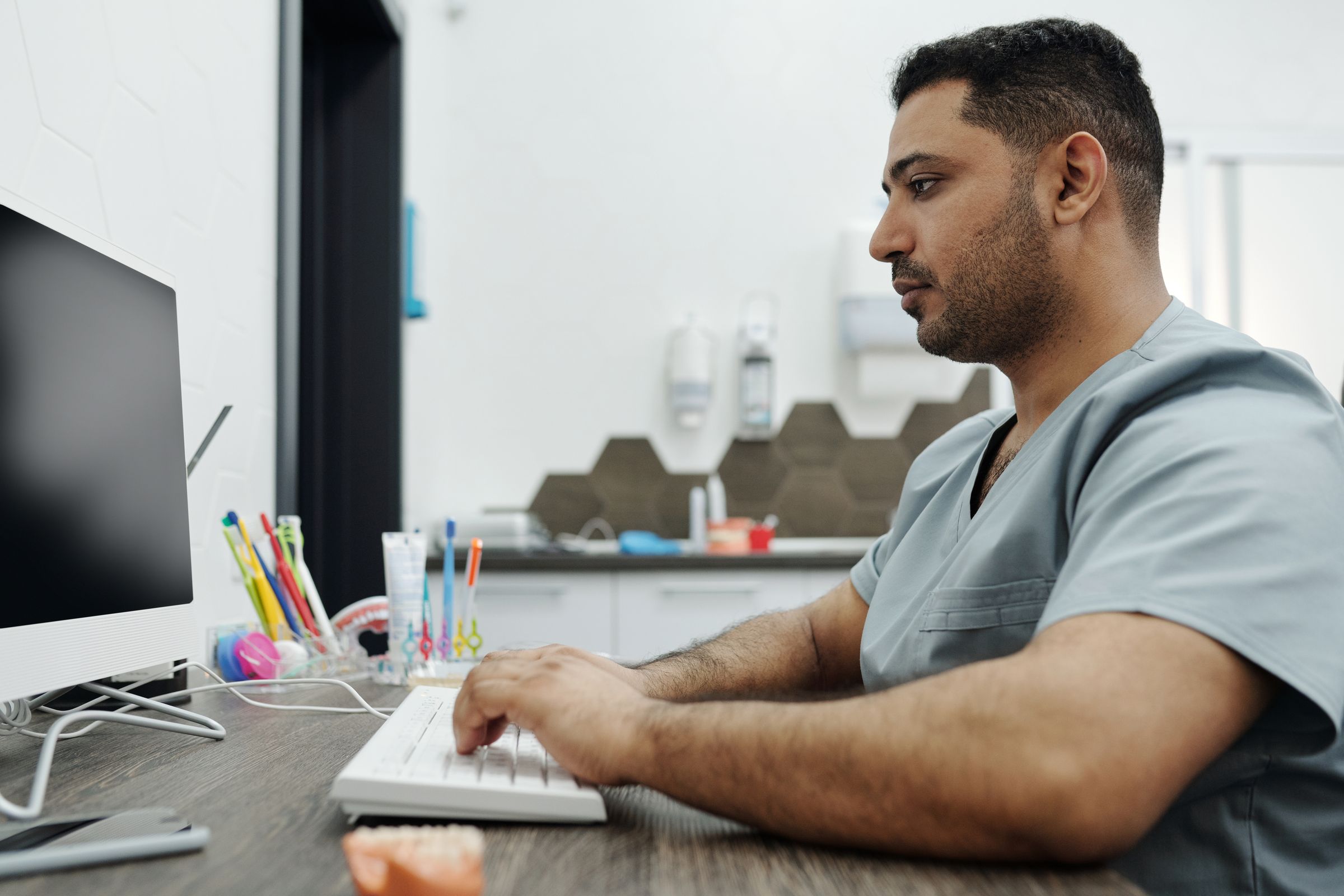 A clinician types at a monitor on a warm wooden worktop, with notes and a keyboard in the foreground.