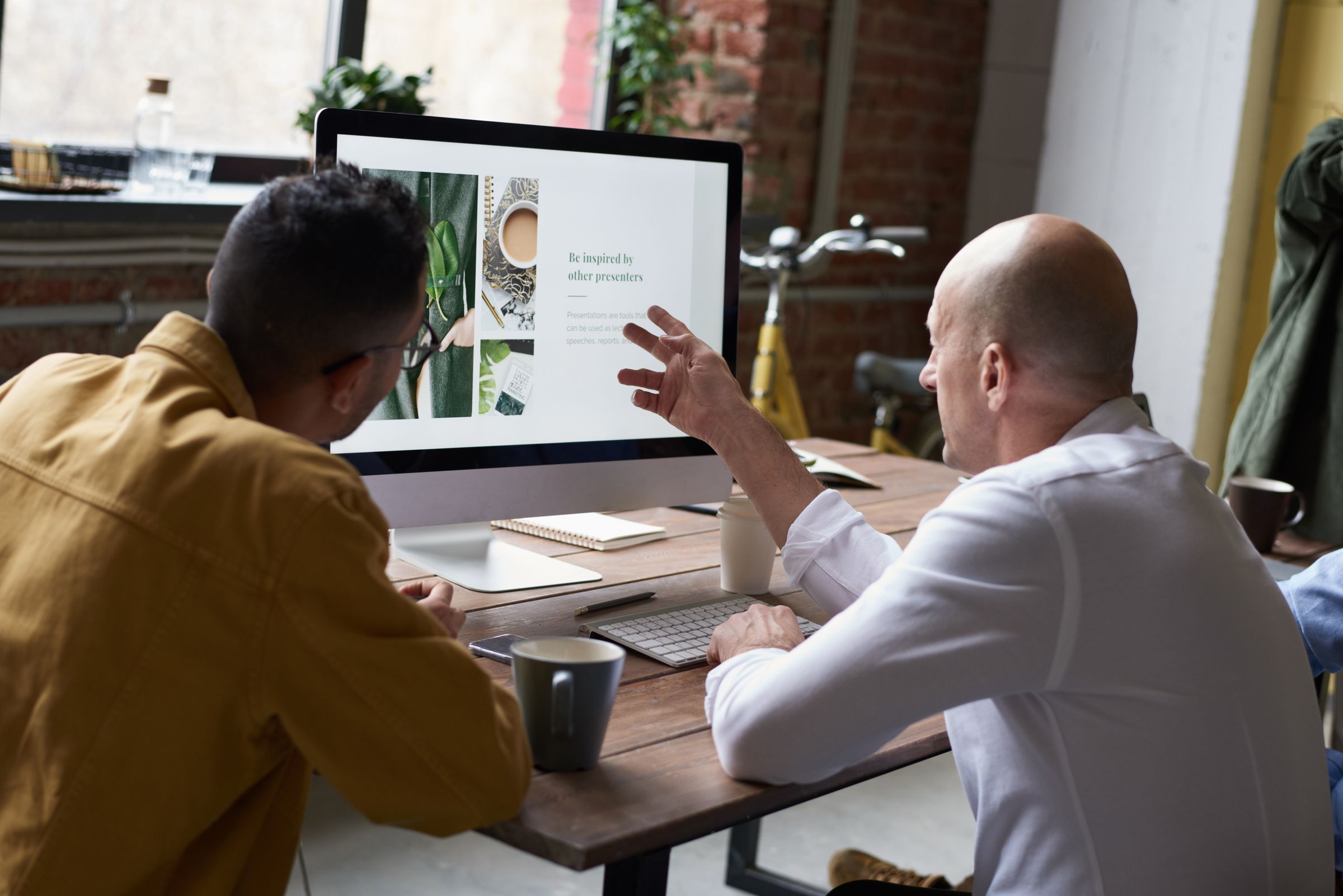Two colleagues sit at a wooden table facing a desktop monitor, one gesturing towards the screen.