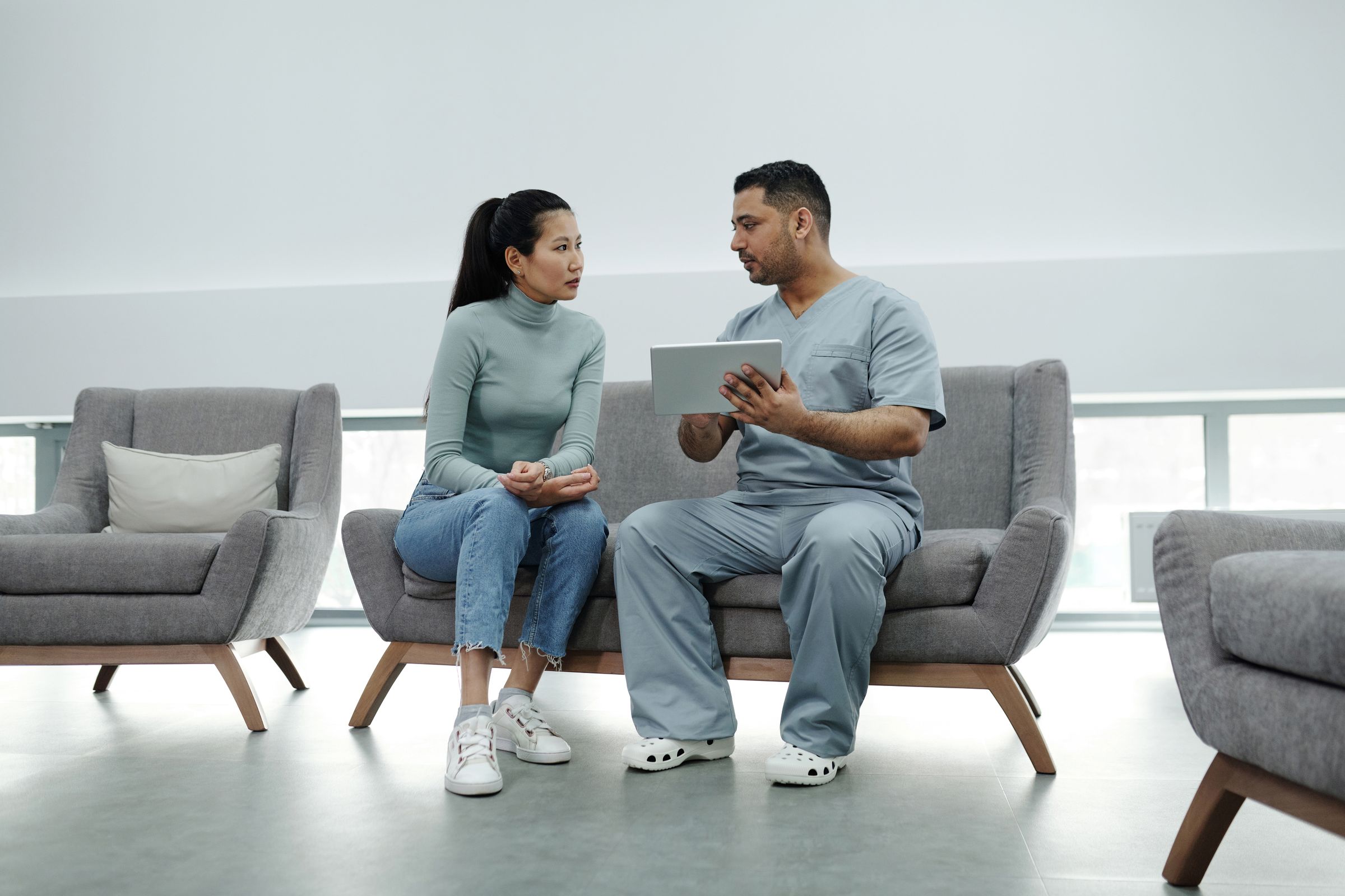 A clinician holding a tablet talks with a seated woman in an airy consulting room.