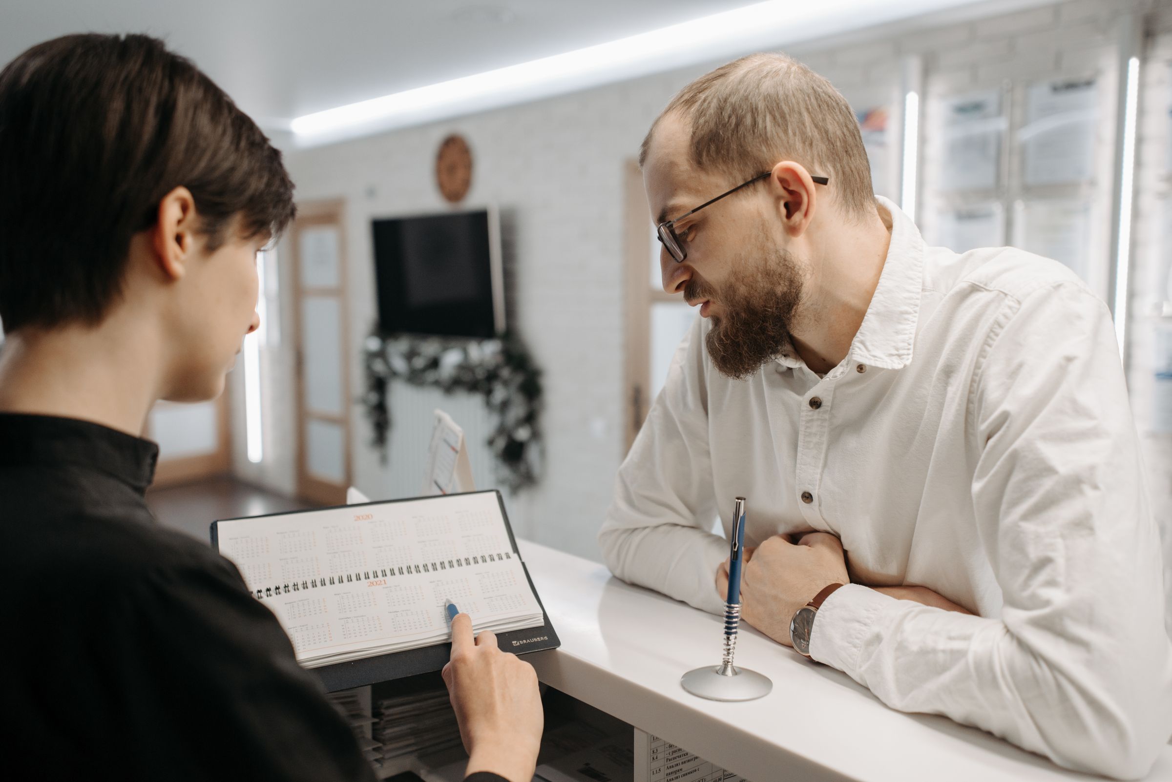 A receptionist turns the pages of a spiral-bound appointment diary resting on the front desk while a man in a white shirt leans in from the other side of the counter to look at it.