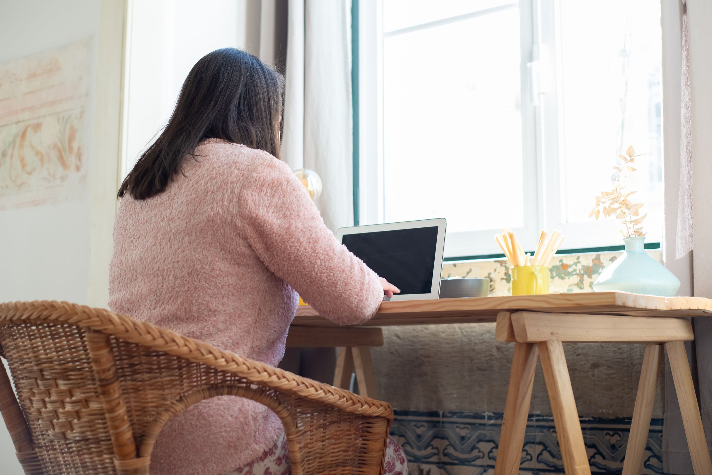 A woman works at a laptop in strong window light, seen from behind.