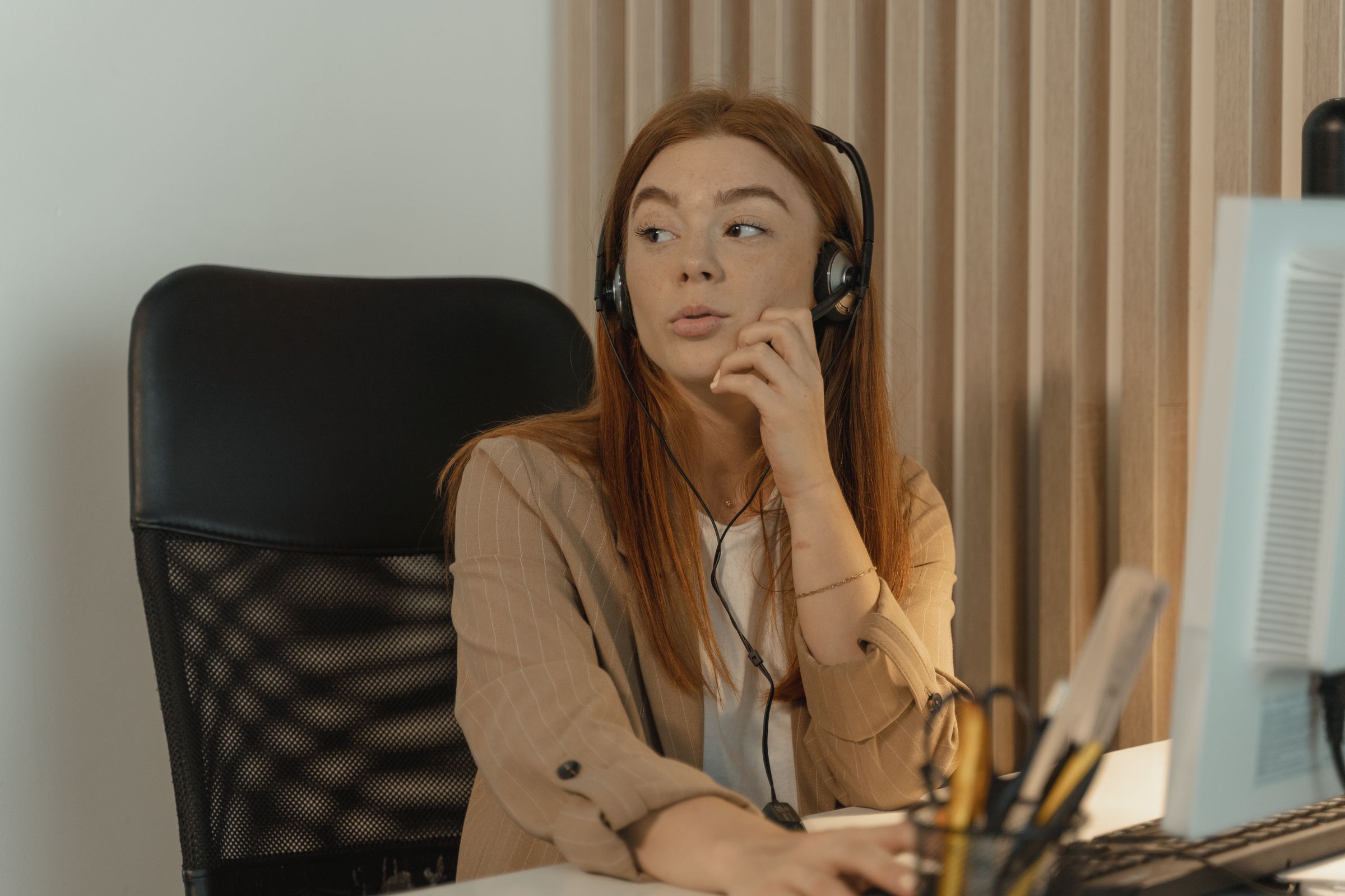 A woman in a tan blazer takes a call on a headset at her desk, with a timber slat wall behind her.